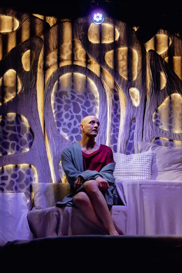 Szenenfoto: Drew Sarich sitzend auf dem Bett mit nachdenklichem Blick im Musical Amanda vor beleuchteter Kulisse.